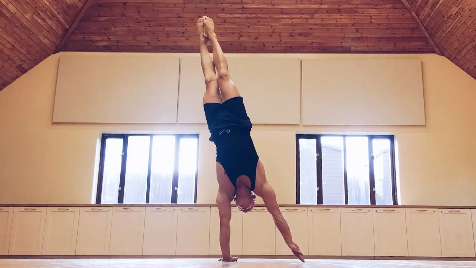 Ben Lowrey in a straight handstand in a warm hall under a timber ceiling, between two bright windows