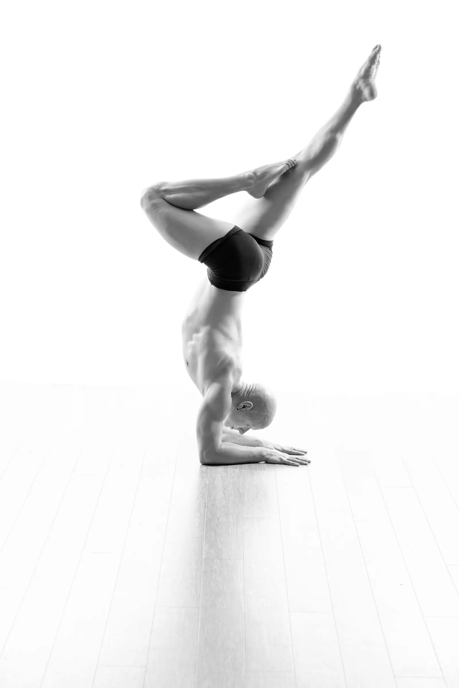 Black-and-white studio photo of Ben Lowrey holding a forearm stand with one knee bent on a white background