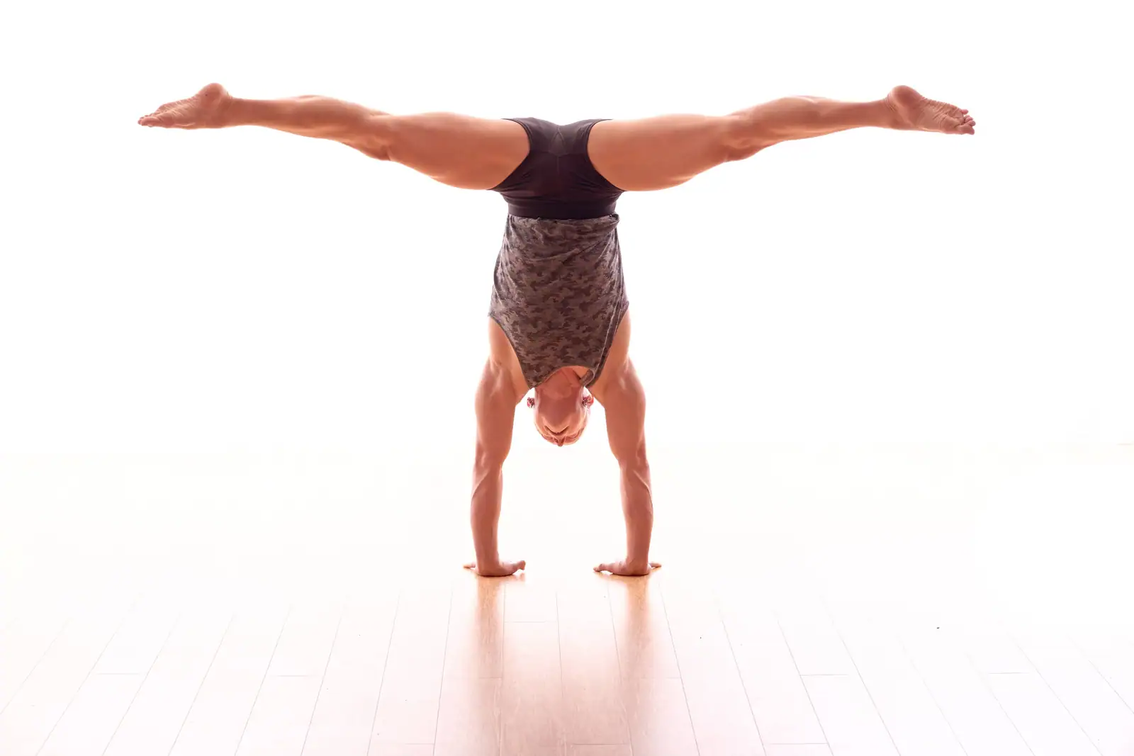 Ben Lowrey in a wide straddle handstand on a bright white studio floor, wearing a camouflage vest