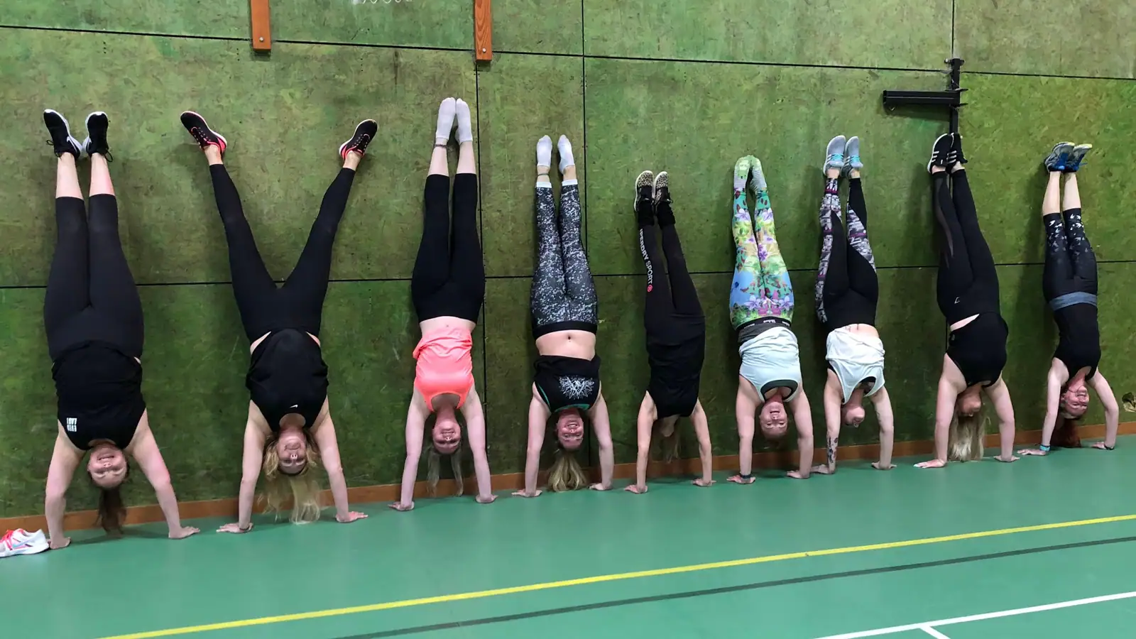 Nine women holding wall handstands in a row against a green sports hall wall