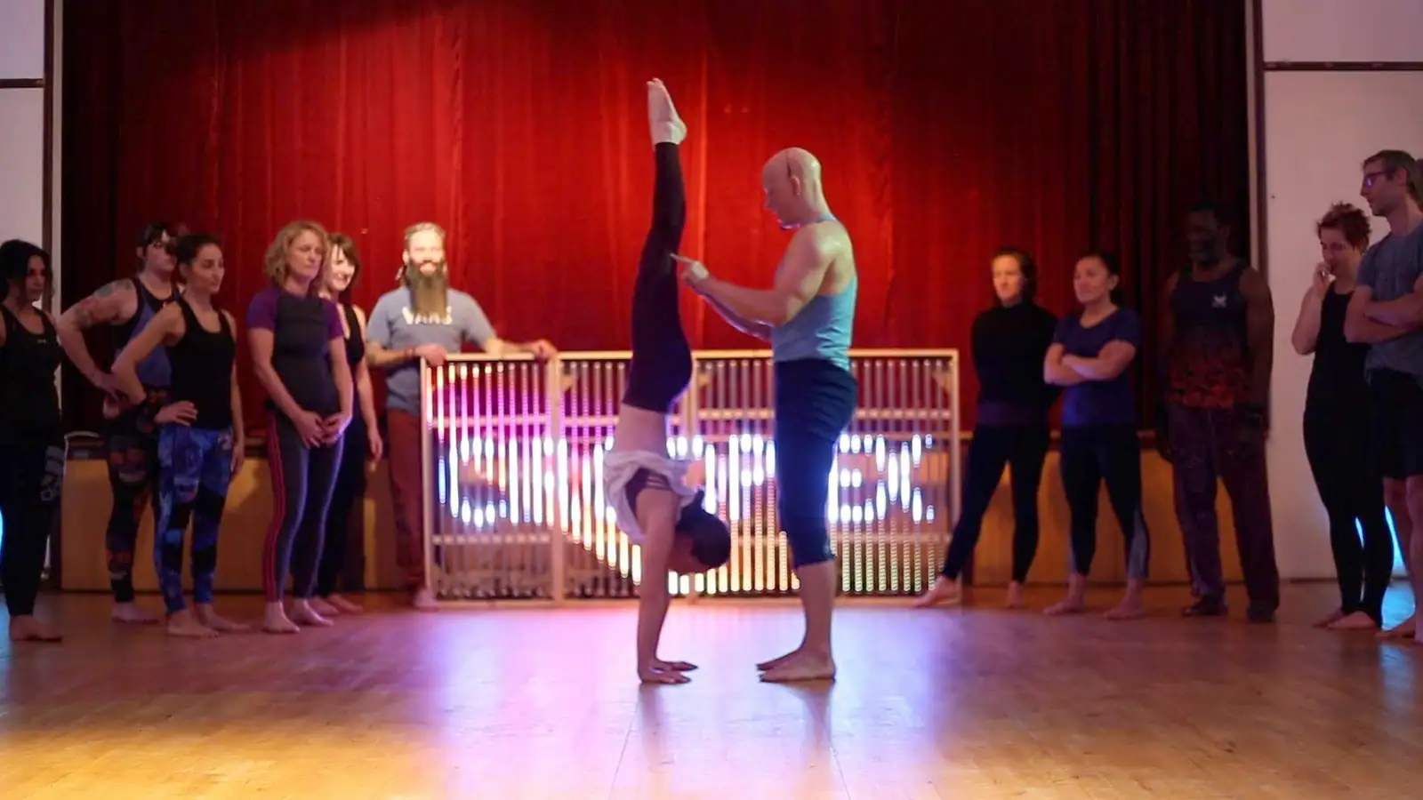 Ben Lowrey spotting a student's handstand in front of the CIRCOPLEX LED sign as a smiling crowd watches